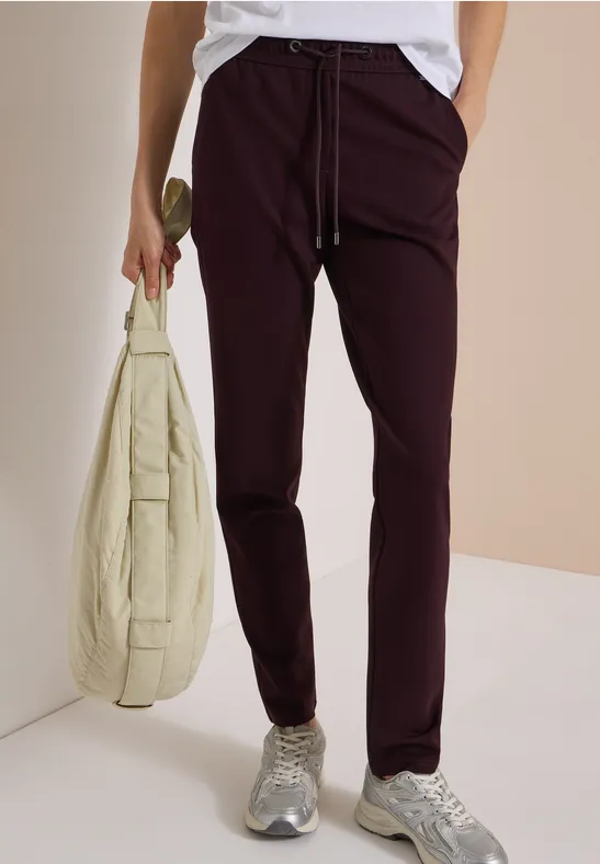 Une personne porte un chemisier blanc, un pantalon bordeaux, des baskets grises et un sac beige.