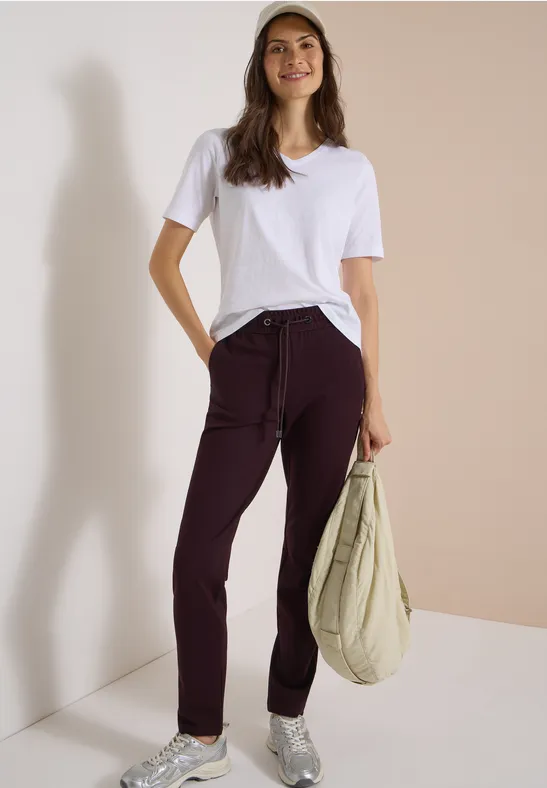 Femme en t-shirt blanc, pantalon bordeaux, casquette et sac beige devant un mur beige clair.
