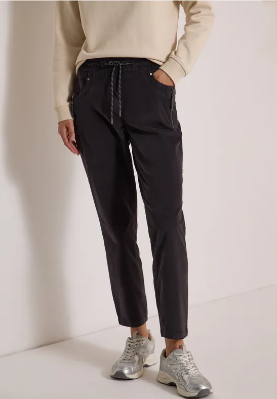 Schwarze Hose mit Tunnelzug, schmaler Schnitt, kombiniert mit hellbeigem Pullover und grauen Sneakern.
