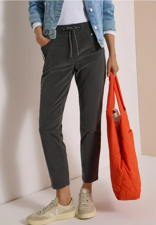 Frau in schwarzer Hose, Jeansjacke, weißen Sneakern und Top, mit orangefarbenem Stepp-Shopper.