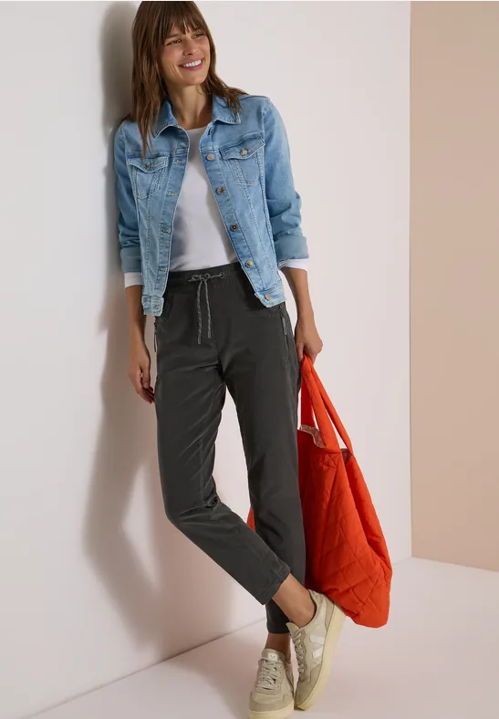 Frau in Jeansjacke, grauem Shirt und schwarzen Jogginghosen lächelt, hält rote Tasche.