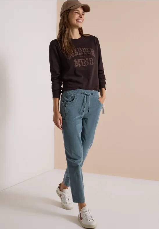 Frau mit Cap trägt braunes Sweatshirt, blaue Chinohose und weiße Sneaker, lässig vor hellem Hintergrund.