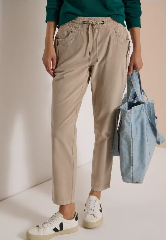 Beige Bundfaltenhose, weiße Sneaker und blaue Stofftasche an der Hand.