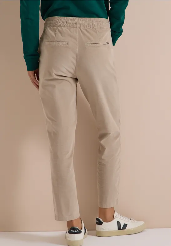 Rückansicht einer Person in beiger Hose und weißen Sneakers mit V-Logo vor neutralem Hintergrund.