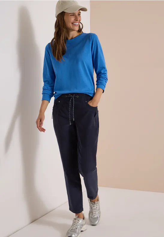 Frau lehnt entspannt an Wand, trägt blaues Sweatshirt, dunkelblaue Hose, weiße Sneaker und weiße Cap.