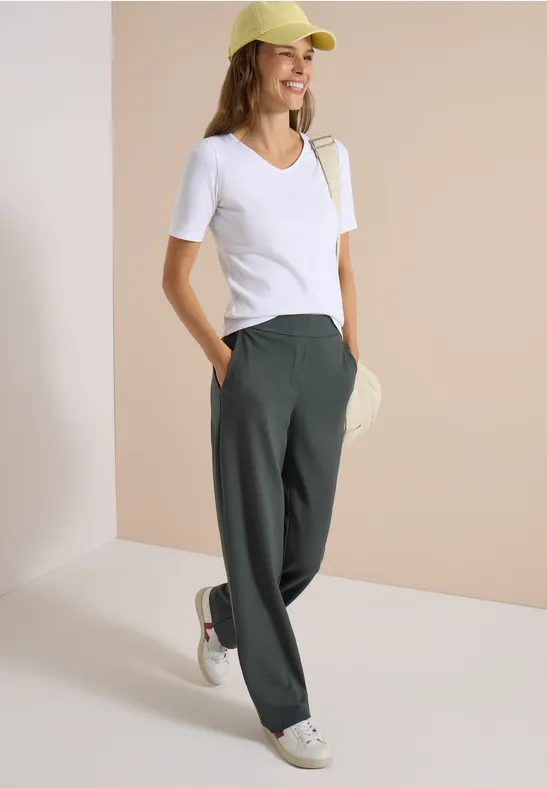 Jeune femme en tee-shirt blanc, pantalon large vert, baskets blanches et casquette jaune devant un mur beige.