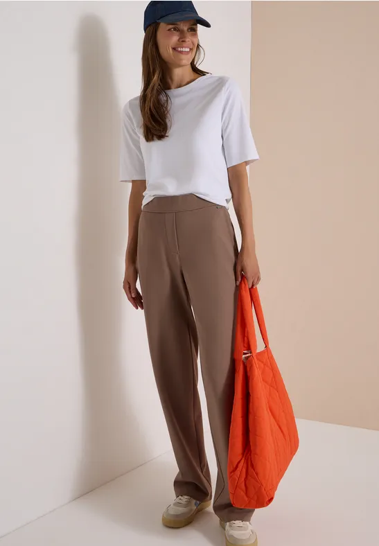 Une femme porte un t-shirt blanc, un pantalon marron, une casquette bleue et un grand sac orange.