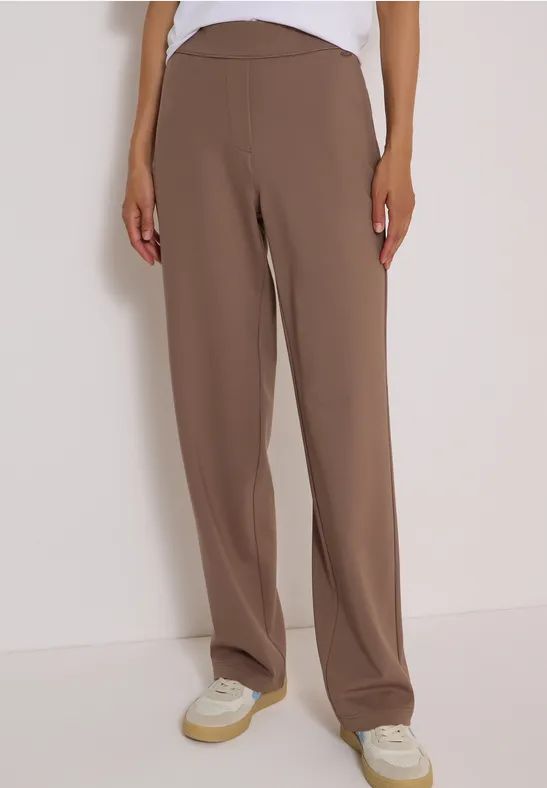 Personne en pantalon marron ample coupe droite et baskets blanches sur fond clair.