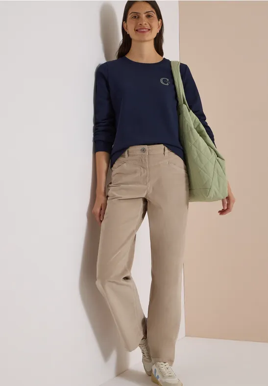 Femme en pull bleu marine, pantalon beige, baskets blanches et sac vert, style casual.