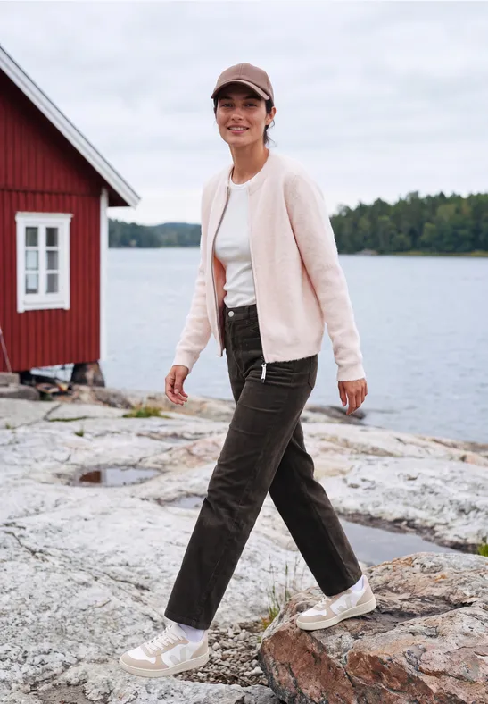 Frau in Jacke und Hose spaziert auf Felsen am See, rotes Haus im Hintergrund