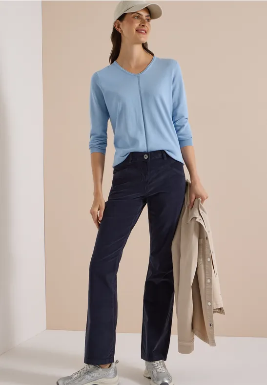 Femme en top bleu clair à col V, pantalon velours côtelé bleu foncé, manteau beige à la main et casquette.