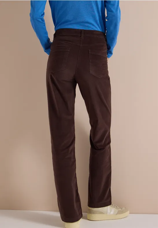 Rückansicht einer Person mit dunkelbraunen Cordhose, blauem Oberteil und hellen Sneakers vor neutralem Hintergrund.