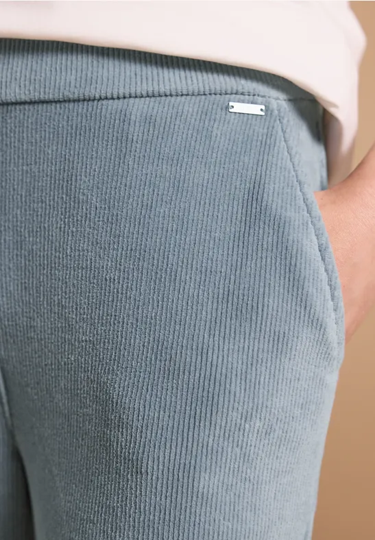 Gros plan d’un pantalon gris côtelé avec poche et petit logo métallique.
