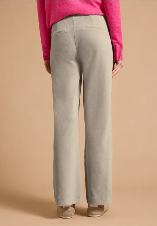 Vue arrière d’une personne portant un pantalon beige ample, un haut rose et des chaussures beiges sur fond uni.