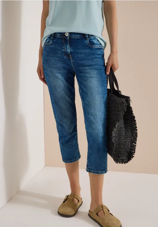 Een persoon draagt blauwe capri-jeans, kaki sandalen en houdt een zwarte gevlochten tas vast.