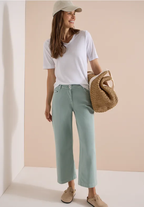 Een vrouw in wit T-shirt, mintgroene culottes, beige pet en strooien tas, glimlachend opzij poserend.