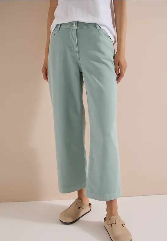 Vrouw draagt wijde pastelgroene broek en beige klompen voor een neutrale achtergrond.