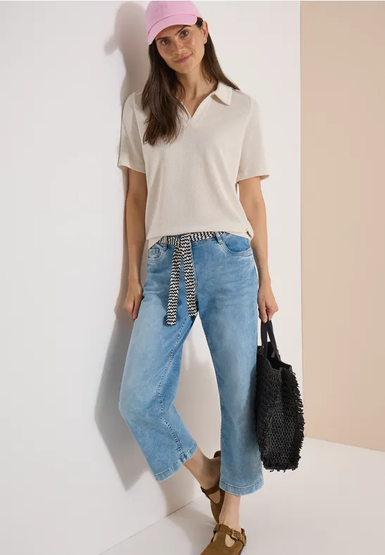 Femme appuyée contre un mur, en jean ample, polo clair, sandales et sac crocheté noir.