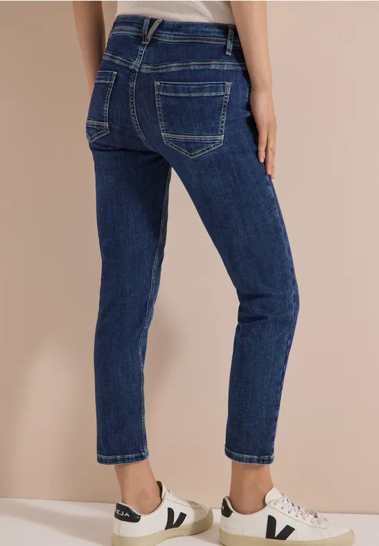 Frau trägt dunkelblaue Jeans und weiße Sneaker, rückseitige Ansicht, beiger Hintergrund.