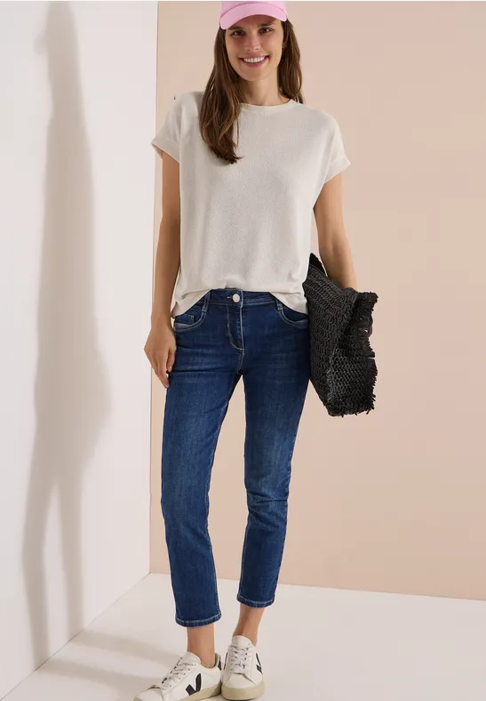 Dame in weißem T-Shirt, blauen Jeans und weißen Sneakern mit schwarzer Flecht-Tasche