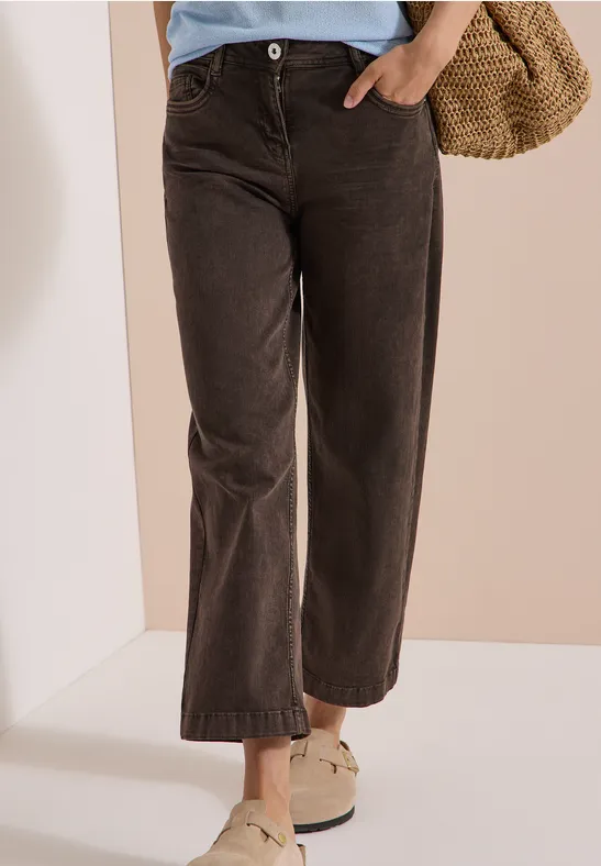 Frau trägt weite dunkelbraune Cordhose mit weißen Slippern und Basttasche.