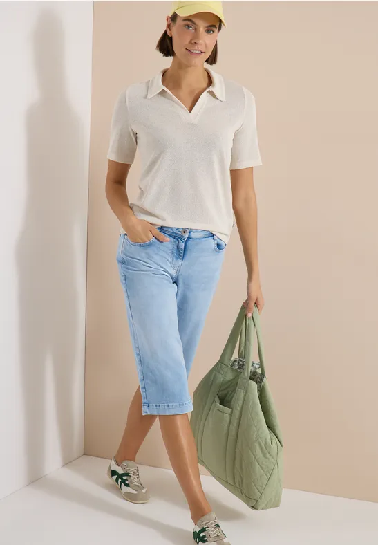 Frau in weißem Polo-Shirt, mittellangem Jeansrock, Sneakern und großer grüner Stepp-Tasche.