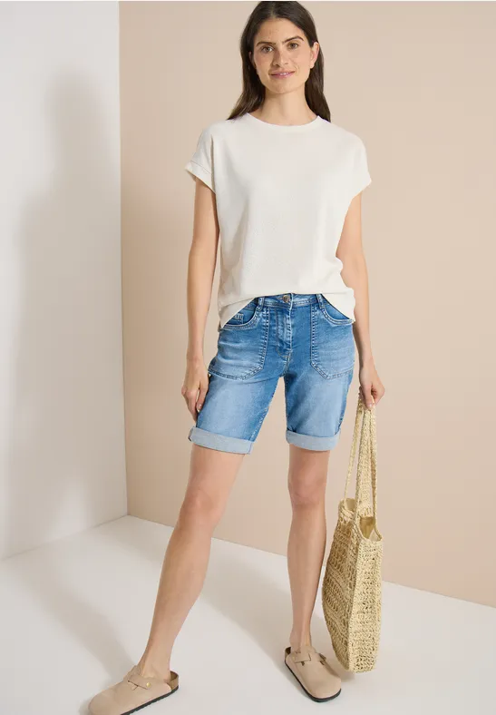 Frau trägt weißes T-Shirt, Jeans-Shorts, Basttasche und Sandalen vor neutralem Hintergrund.