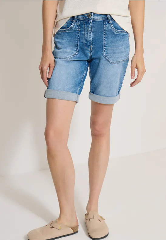 Frau trägt helle Jeans-Shorts und beigefarbene Hausschuhe, steht vor neutralem Hintergrund.