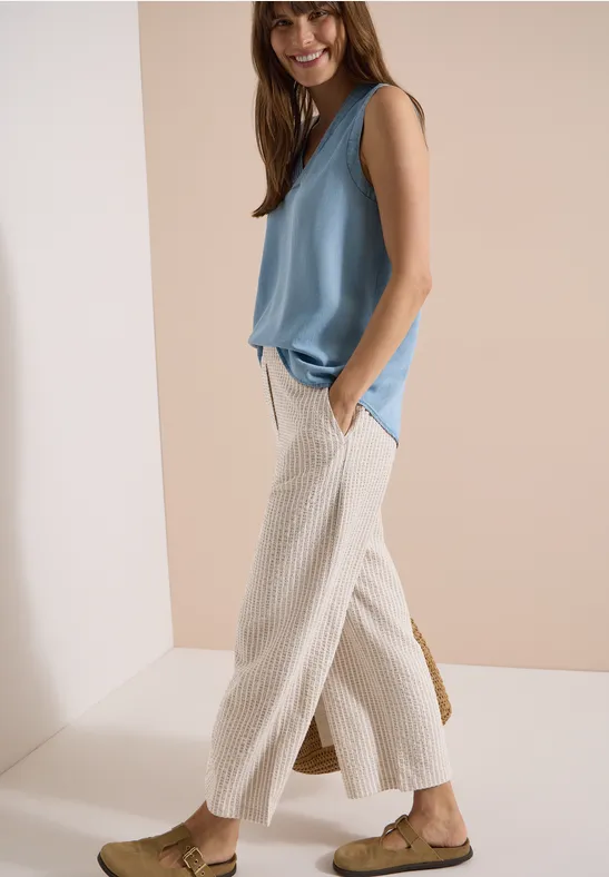 Modell mit ärmelloser hellblauer Bluse, cremeweißer weiter Leinenhose und flachen Sandalen.