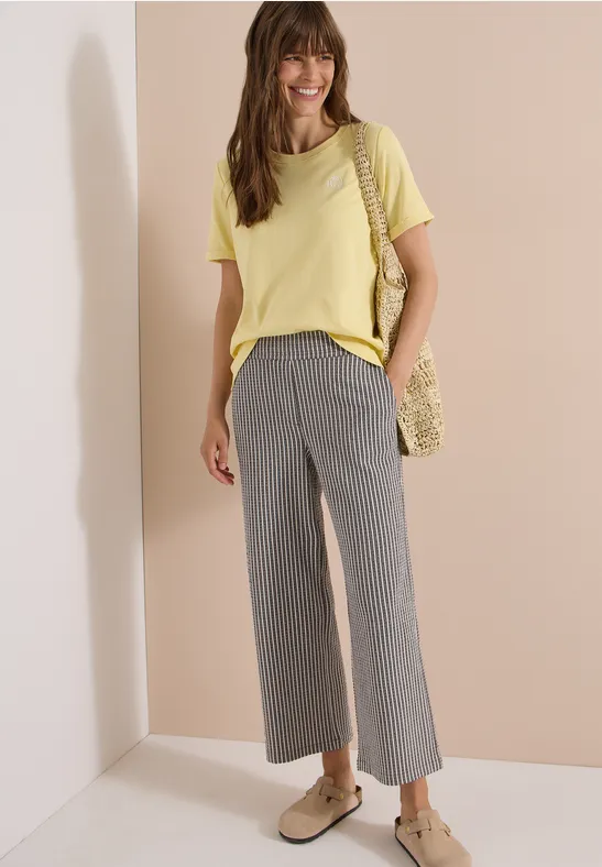 Femme en t-shirt jaune, pantalon large gris, chaussures plates et sac en crochet jaune.