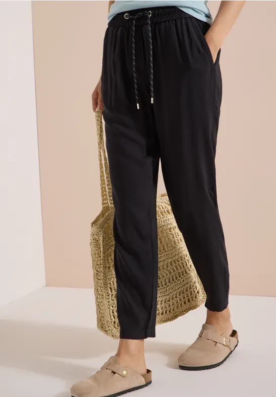 Schwarze, lässige Stoffhose mit Tunnelzug, kombiniert mit Basttasche und beigen Slippern.