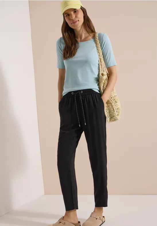 Frau in hellblauem Shirt und schwarzen Kordelhosen mit Strohtasche und Sandalen vor schlichtem Hintergrund.