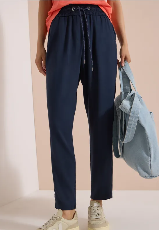 Femme en pantalon bleu marine à cordon, baskets blanches, tenant un sac en jean bleu clair.