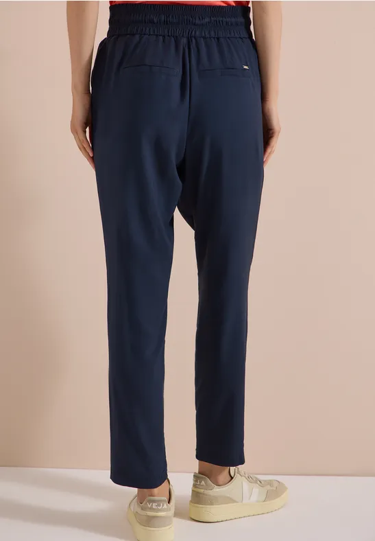 Vue arrière d'une personne portant un pantalon de jogging bleu marine, taille élastique, et baskets blanches devant un mur clair