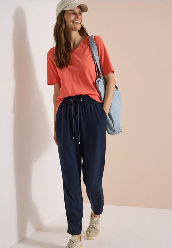 Femme en t-shirt rouge, pantalon de jogging bleu marine et baskets beiges, avec un sac en jean.