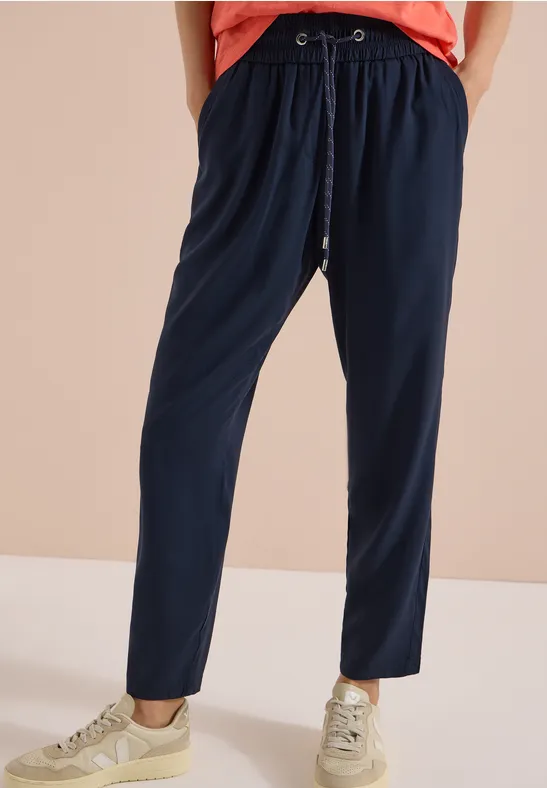 Jambes portant un pantalon de jogging bleu marine à cordon et des baskets claires sur fond beige.