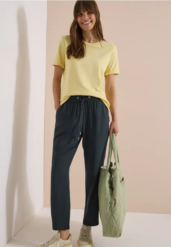 Frau trägt gelbes T-Shirt, schwarze Hose und hält eine grüne Tasche vor beigem Hintergrund.