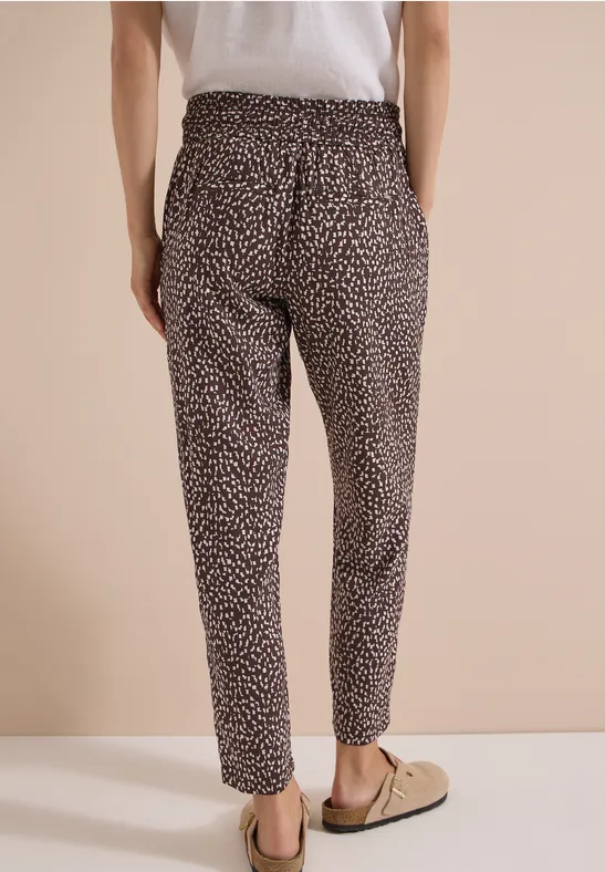 Abgeschnittenes Bild von einer Person in leopardgemusterten Stoffhosen und Slip-on-Schuhen vor beigem Hintergrund.