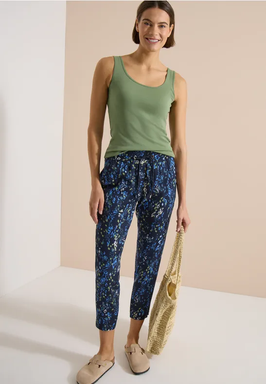 Eine Frau trägt ein grünes Top, gemusterte blaue Hosen und beige Slipper; sie hält eine Tasche.