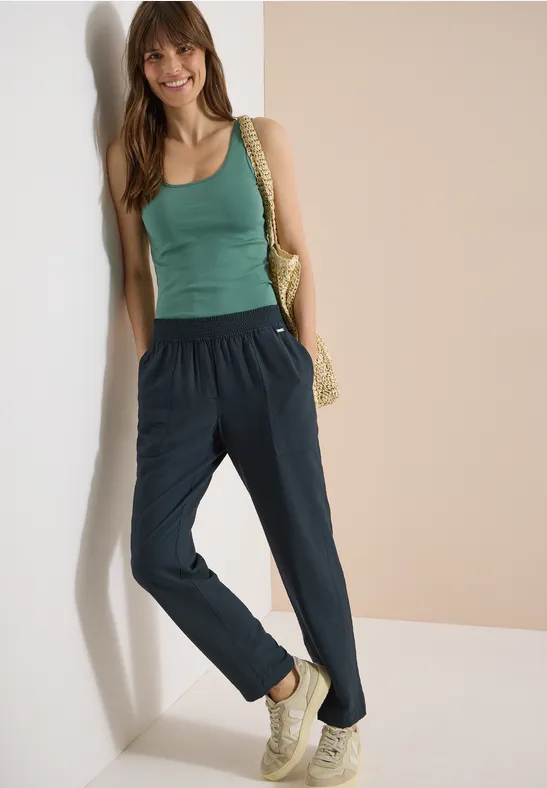 Vrouw leunt tegen muur, draagt groene top, blauwe broek, sneakers en strozak.