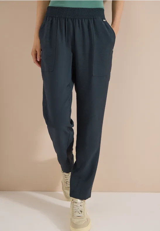 Losse donkerblauwe broek met zakken en witte sneakers, gedragen tegen een effen beige achtergrond.