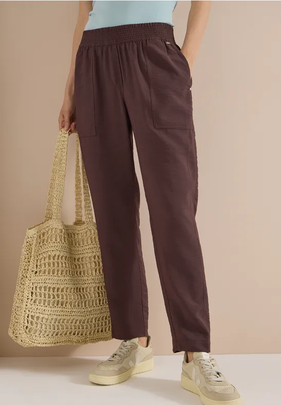 Vrouw met bruine, comfortabele broek, crème top, beige sneakers en gehaakte tas.