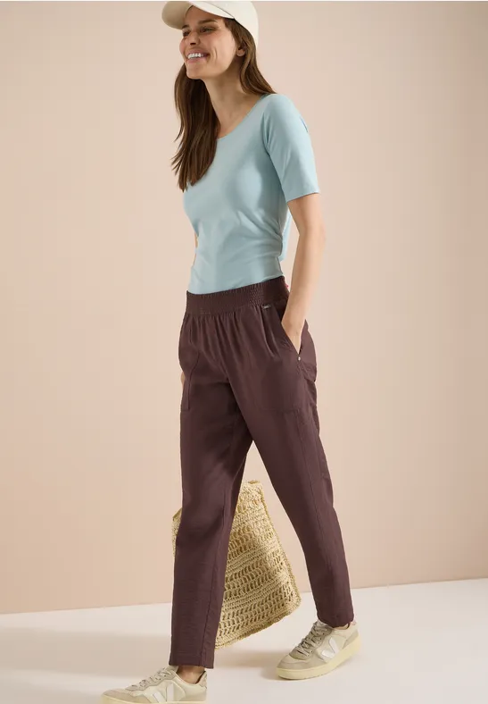 Vrouw in lichtblauw T-shirt, donkerrode vrijetijdsbroek en strooien tas, ontspannen pose.