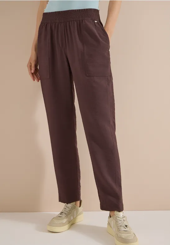 Een vrouw draagt een donkerbruine, losse broek met zakken en witte sneakers voor een beige achtergrond.