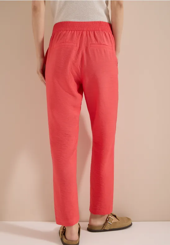 Iemand van achteren voor een beige muur met een koraalrode broek en groene slippers.