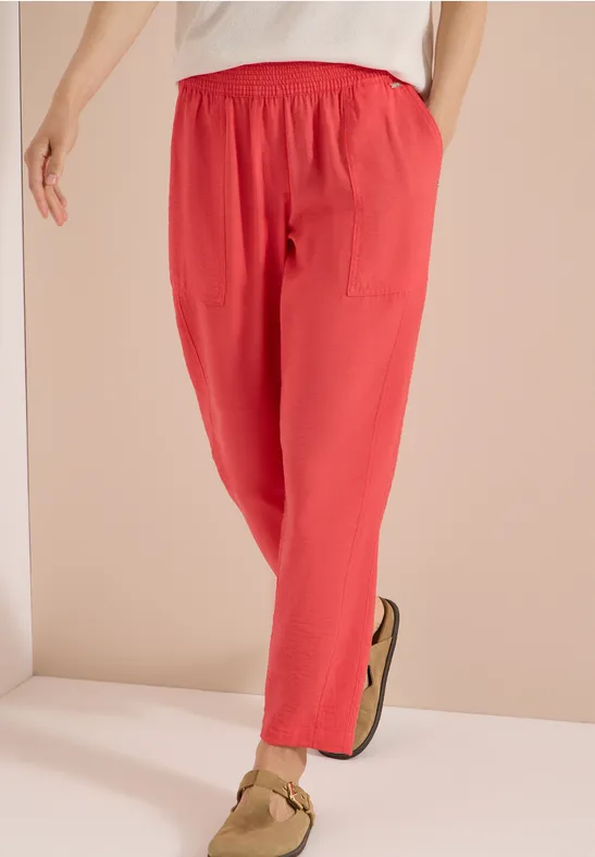 Persoon draagt een losse koraalkleurige linnen broek met elastische taille en zijzakken, gecombineerd met beige sandalen.