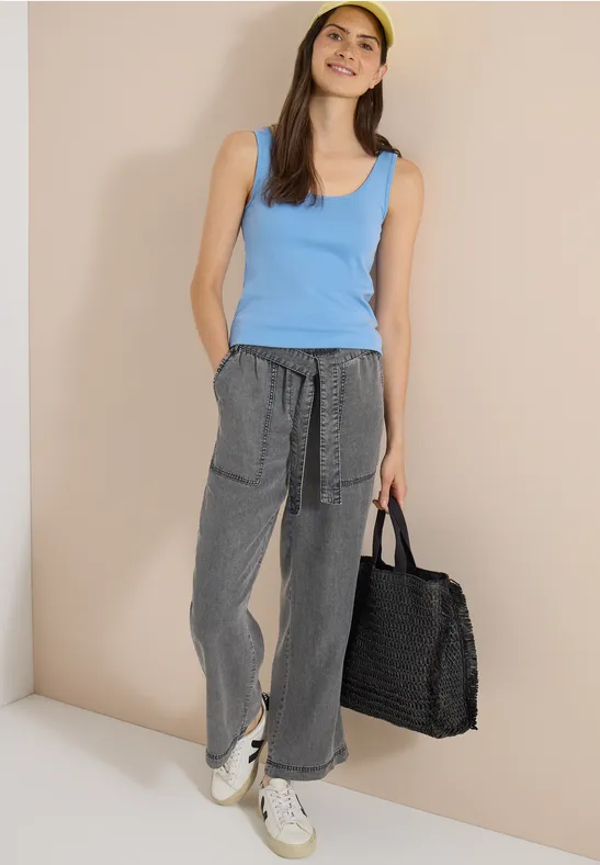 Femme en débardeur bleu, pantalon ample gris, baskets blanches, portant un grand sac en osier noir.