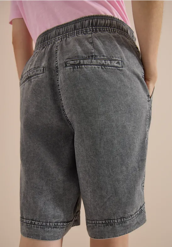 Eine Person trägt graue Denim-Bermuda-Shorts mit Gummibund, Gesäß- und Seitentaschen, steht mit Händen in den Taschen.