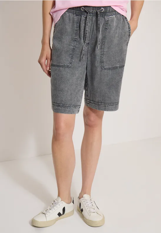 Person mit grauen Jeans-Shorts, Taschen und weißen Sneakern vor neutralem Hintergrund.