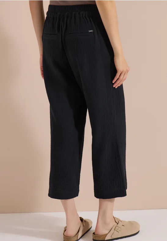 Rückenansicht: schwarze, weite, knöchellange Rippstrick-Hose mit elastischem Bund und hellen Slippers.